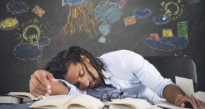 Teacher lays head on arm on desk with colorful chalkboard behind him