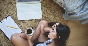Looking down on girl sitting on floor as she holds coffee cup and puts head in hands in front of computer