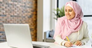 Young woman with hijab smiles at computer and writes something down at desk